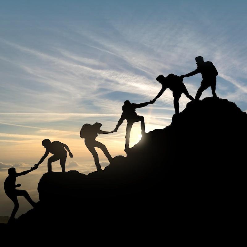 Silhouetted view of people helping each other up a slope to the top of a rocky hill